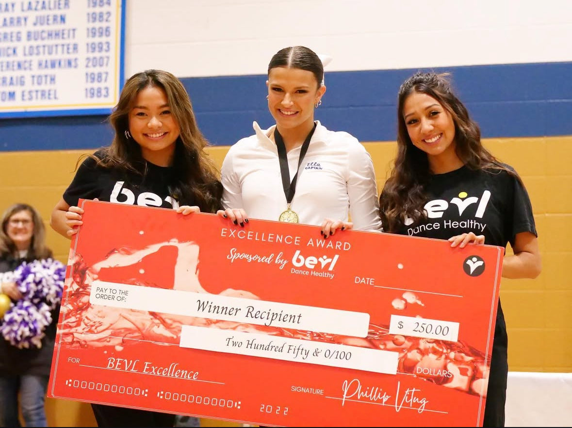 Three women holding an excellence award check in a gymnasium.
